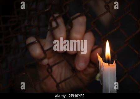 old female hands hang on a fence made of iron mesh in the lock on the background of a burning candle Stock Photo