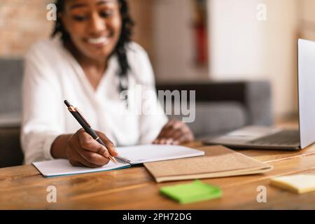 Happy black woman writing in notepad and using laptop, noting information from internet, selective focus Stock Photo