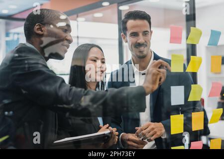 Positive diverse coworkers in formal clothes standing near glass wall with colorful sticky notes in office and discussing project Stock Photo