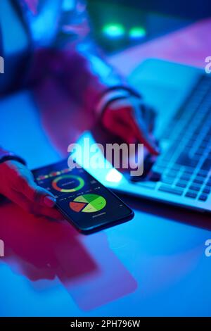 Neon metaverse futuristic concept. Closeup on woman using laptop, using smartphone and exploring graphs in office. Stock Photo