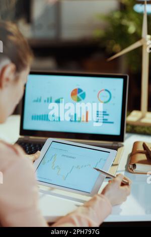 Seen from behind modern woman with laptop working with charts, using digital tablet and analysing investment marketing data in modern office. Stock Photo