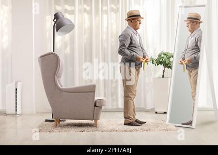 Elderly man measuring his waist and standing in front of a mirror at home Stock Photo