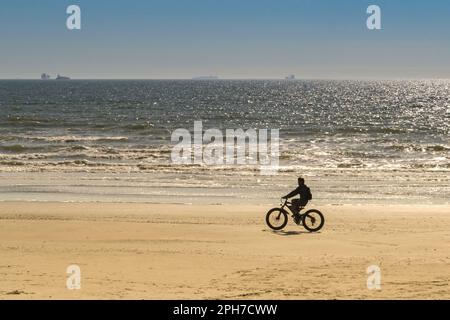 Galveston, Texas, USA - February 2023: Silhouette of a person riding an electric bicycle across the sand on the city's beach at low tide. Stock Photo