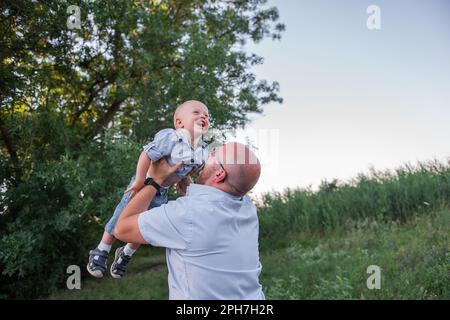 Bald man with glasses throws child into the sky air. Father in jeans plays, embrace with son in nature outside the city. Little boy laughs, having fun Stock Photo