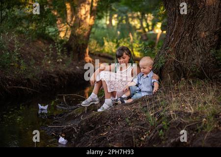 Girl and boy are sitting on the bank of river, launching white paper origami boat into the water. Teenage sister with green hair hugs brother toddler. Stock Photo