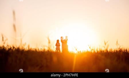 Mothers silhouette with little son in arms and daughter in sunset on the field. Orange natural background. Panoramic view. Traveling with children to Stock Photo