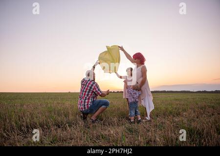 Diversity family launch yellow sky lantern in field at sunset. Mother, father holding Chinese paper lantern, daughter, son wave hands. Making wishes f Stock Photo