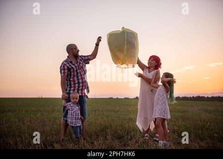 Diversity family launch yellow sky lantern in field at sunset. Mother, father holding Chinese paper lantern, daughter, son wave hands. Making wishes f Stock Photo