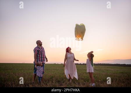 Diversity family launch yellow sky lantern in field at sunset. Mother, father holding Chinese paper lantern, daughter, son wave hands. Making wishes f Stock Photo