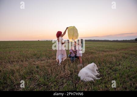 Diversity family with Samoyed dog launch yellow sky lantern in field at sunset. Mother, father holding Chinese paper lantern, daughter, son wave hands Stock Photo