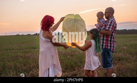 Diversity family launch yellow sky lantern in field at sunset. Mother, father holding Chinese paper lantern, daughter, son wave hands. Making wishes f Stock Photo