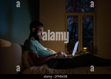 Concentrated African American programmer man lying on bed work on laptop at night.  Stock Photo