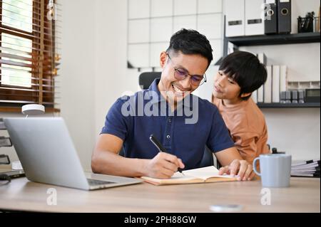 Happy and cheerful millennial Asian dad is being annoyed from his son while working in his home office. Stock Photo