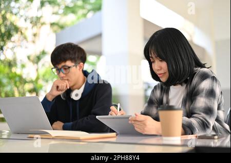 Pretty and smart young Asian female college student working on her schoolwork on her tablet while her male friend works on his homework on his laptop, Stock Photo