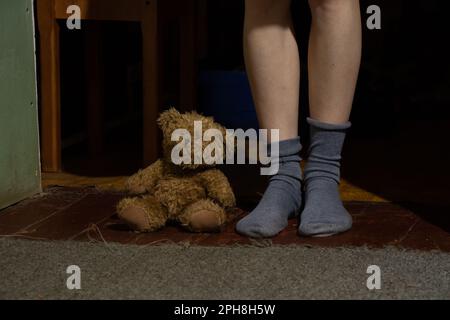 female feet in socks and brown teddy bear on the floor in a dark room Stock Photo