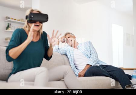 Its so realistic. a mature woman using a VR headset with her husband in the background. Stock Photo