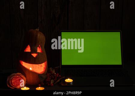 Halloween pumpkins on rough wooden boards with candles and rose glowing in the dark with open laptop with green screen blank space, laptop with green Stock Photo