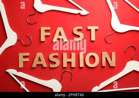 Phrase FAST FASHION made of wooden letters and white hangers on red background, flat lay Stock Photo