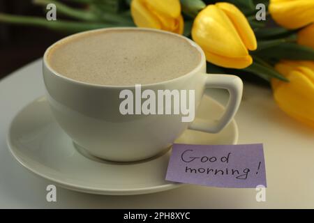 Cup of coffee, flowers and card with phrase GOOD MORNING! on white table, closeup Stock Photo