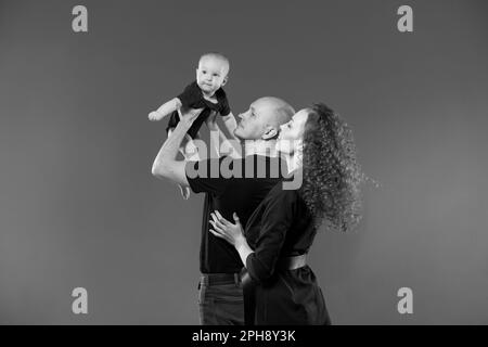 Portrait happy young family, dad holds his son in raised arms, mom stands next to him smiling. Together they are happy posing against the backdrop in the studio. Family concept. Stock Photo