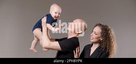 Portrait happy young family, dad holds his son in raised arms, mom stands next to him smiling. Together they are happy posing against the backdrop in the studio. Family concept. Stock Photo