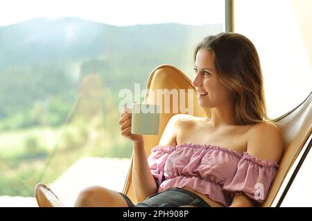 Happy woman sitting on a chair holds coffee cup looking through a window at home Stock Photo