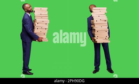 Office employee carrying big pile of pizza packs in studio, holding fast food meal over full body green screen background. Startup businessman in suit having takeout order on camera. Stock Photo