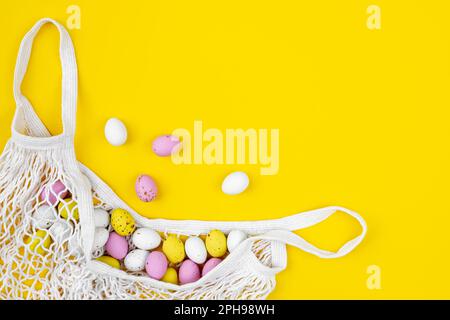 Colored Easter eggs in a mesh bag on a yellow background, flat lay. Stock Photo