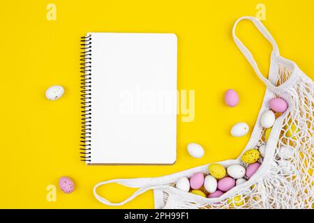 Colored Easter eggs in a mesh bag and a blank notepad on a yellow background. Stock Photo