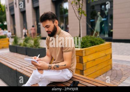 Creative male artist with tattooed hands sitting on bench and making sketches with pen on notebook. Talented young man with pencil and sketchbook Stock Photo