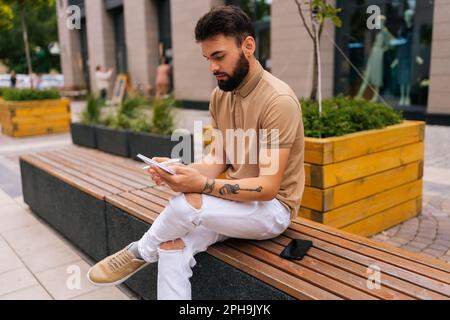 Portrait of handsome male artist with tattooed hands sitting on bench with takeaway coffee and making sketches with pen on copybook. Stock Photo