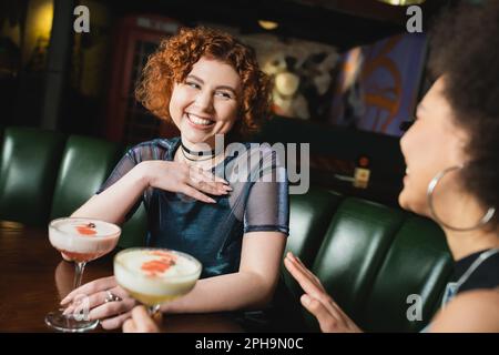 Positive redhead woman holding clover club cocktail near african american friend in bar,stock image Stock Photo