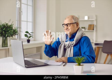 Happy retired senior man waving hand talking to webcam making video call on laptop at home. Stock Photo