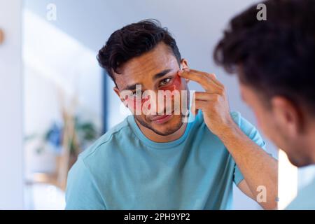 Biracial man looking in mirror and applying under eye masks in bathroom Stock Photo