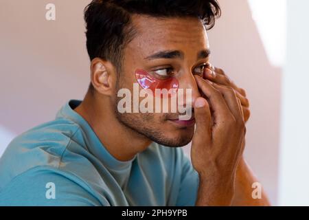 Biracial man looking at mirror and applying under eye masks in bathroom Stock Photo