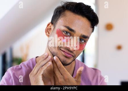 Smiling biracial man with eye masks inspecting his face in bathroom mirror Stock Photo