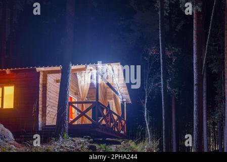 A wooden cottage for relaxing at night glows with garlands in the dark on a mountain in a pine forest with a terrace with curtains in Altai. Stock Photo