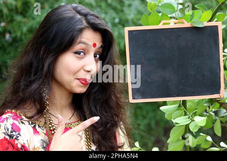 a beautiful smiling girl pointing to a black chalk board for educational, promotional, business, marketing announcement or advertisement. Can be used Stock Photo