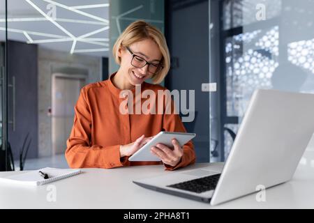 Young successful businesswoman at workplace is satisfied with results of achievement at work smiling and using tablet computer, blonde programmer is testing new software in application. Stock Photo