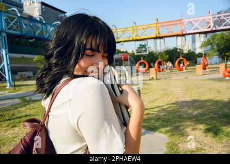 young happy latin woman, venezuelan university student outdoors with closed eyes hugging a book, school concept, copy space. Stock Photo