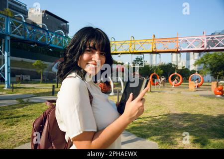 happy young latin venezuelan student outdoors standing in profile, holding a book with her hands, smiling and looking at the camera. school concept, c Stock Photo