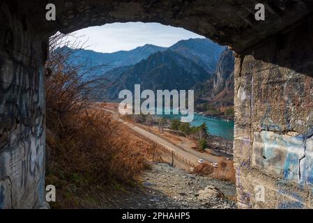 The exit from the old abandoned adit of the cave among the stones glows with daylight in the dark in Altai against the backdrop of the river and mount Stock Photo