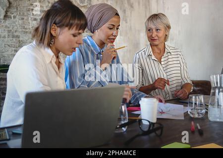 Three Caucasian women collaborate in an office, one wearing a Middle Eastern turban. They discuss a project around a desk with laptops.' Stock Photo