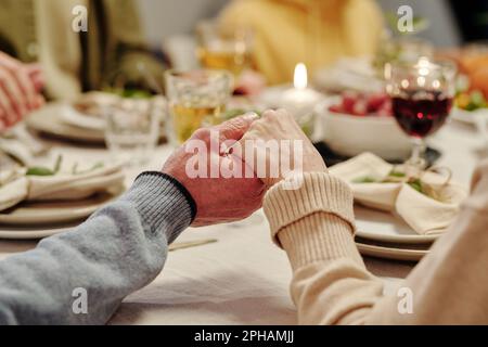 Hands of affectionate senior couple praying by served festive table with homemade food and burning candles before Thanksgiving dinner Stock Photo