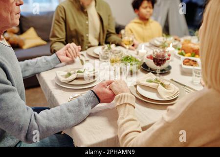 Senior couple holding by hands by festive table served with homemade food and beverages while praying before family dinner Stock Photo