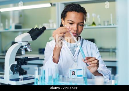 Young female pharmacologist with dropper taking sample of blue liquid substance from flask during clinical experiment in laboratory Stock Photo