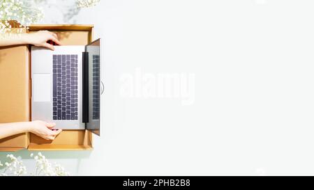 Buying and delivering new laptop. Romantic overhead shot of womans hands holding laptop in cardboard box on blue background with white spring flowers. Stock Photo