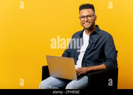 Nice smiling indian male entrepreneur using laptop sitting on the armchair isolated on yellow, portrait of happy determined male student looking at camera with computer on his lap, arab man typing Stock Photo