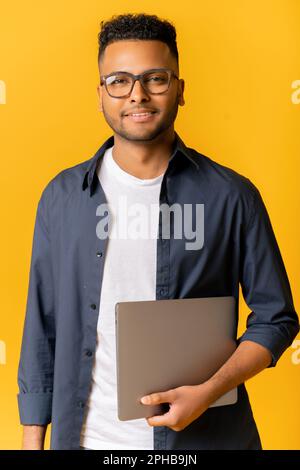 Portrait of successful indian man in glasses standing and carrying laptop, looking at the camera isolated on yellow, cheerful male entrepreneur, freelancer or student smiling Stock Photo