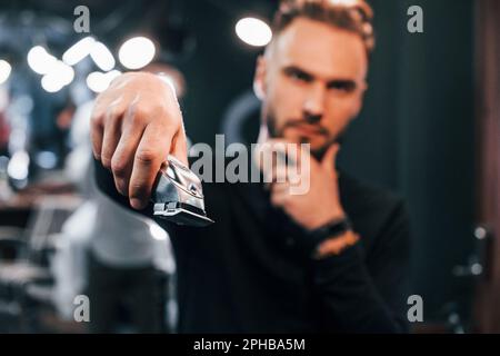 Young bearded man standing in barber shop and holding clipper Stock Photo